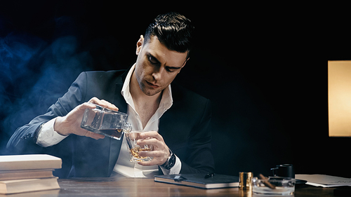 Businessman pouring whiskey near books and cigar on black background with smoke