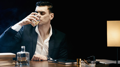 Businessman in suit drinking whiskey near books and ashtray on table isolated on black