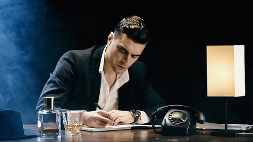 Businessman writing on notebook near whiskey and telephone on black background with smoke