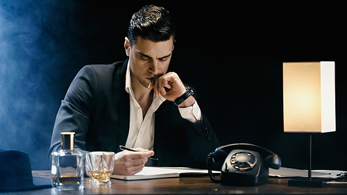 Businessman looking at notebook near telephone and whiskey on table on black background with smoke