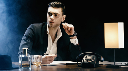 Businessman looking away near notebook and whiskey on table on black background with smoke