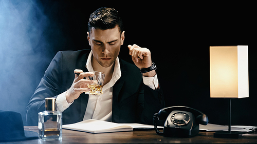 Businessman holding whiskey near notebook and telephone on black background with smoke