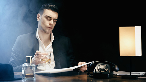 Businessman holding whiskey and notebook near telephone and lamp on black background with smoke
