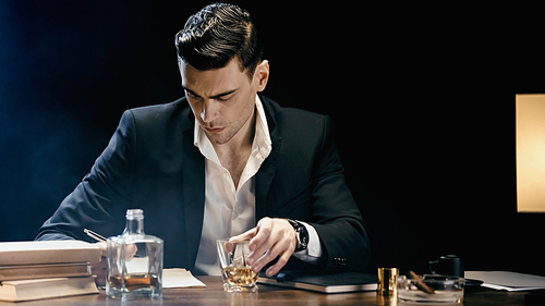 Businessman holding glass of whiskey near books and ashtray on table on black