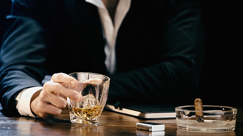 Cropped view of businessman holding glass of whiskey near cigar in ashtray isolated on black