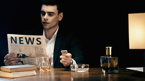 Blurred businessman reading newspaper and holding lighter near ashtray and whiskey on table isolated on black
