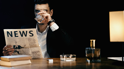 Businessman drinking whiskey and reading newspaper near books and ashtray on table isolated on black