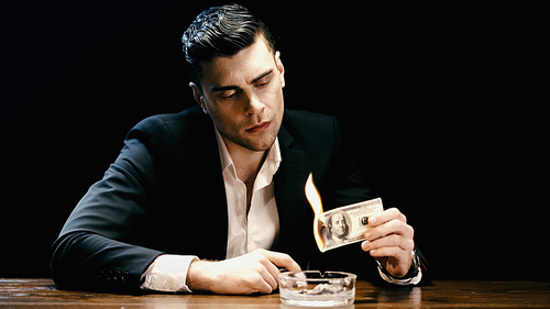 Businessman in suit holding burning dollar banknote near ashtray isolated on black