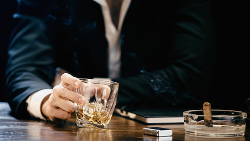 Cropped view of blurred businessman holding glass of whiskey near cigar in ashtray isolated on black