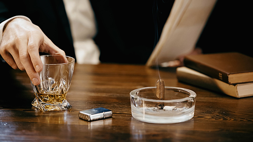 Cropped view of businessman holding glass of whiskey near cigar with smoke in ashtray isolated on black