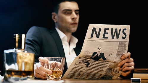 Blurred businessman reading newspaper and holding glass of whiskey isolated on black