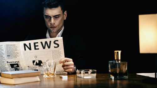 Businessman reading newspaper near whiskey and books on table isolated on black