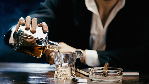 Cropped view of blurred businessman pouring whiskey near cigar in ashtray on black