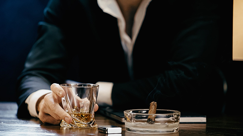 Cropped view of businessman holding glass of whiskey near cigar and lighter isolated on black