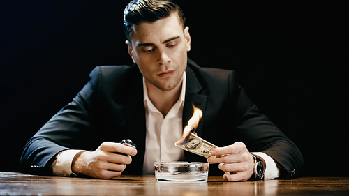 Businessman holding burning dollar and lighter near ashtray on table isolated on black