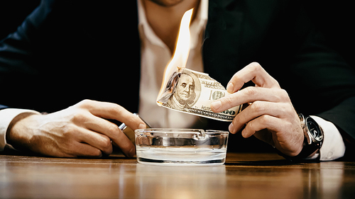 Cropped view of businessman holding burning money and lighter near ashtray isolated on black