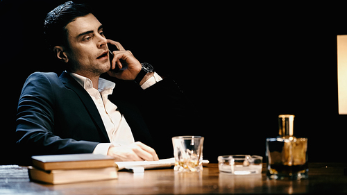 Businessman talking on smartphone near whiskey and books on table isolated on black