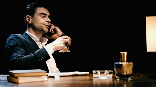Businessman talking on smartphone and holding whiskey near books and newspaper on table isolated on black
