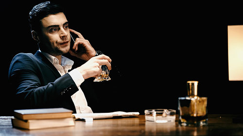displeased businessman holding whiskey and talking on cellphone near books and bottle isolated on black