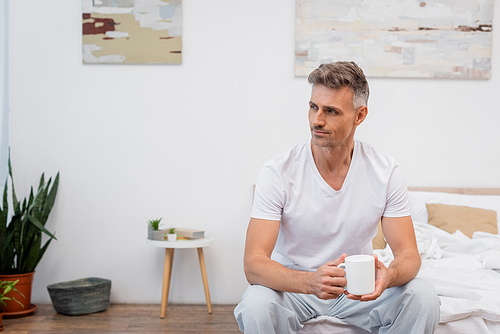 Man in pajamas holding cup of coffee on bed at home