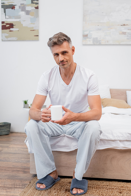 Man in pajamas and slippers holding cup of coffee while sitting on bed