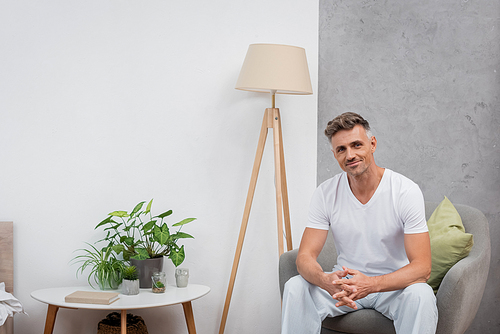 Smiling man in pajamas looking at camera while sitting on armchair at home