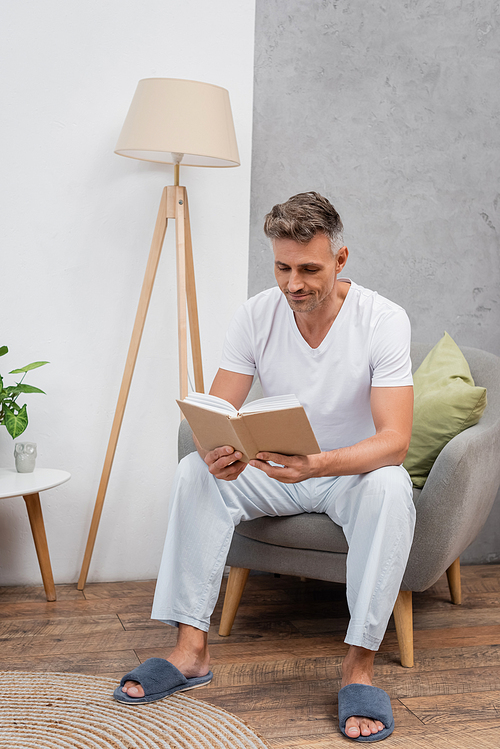 Man in pajamas and slippers reading book on armchair at home