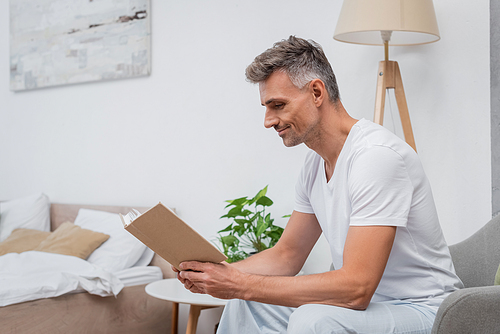 Side view of man in pajamas reading book on armchair in bedroom