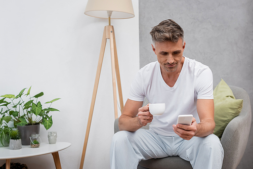 Man holding cup of coffee and using smartphone on armchair at home