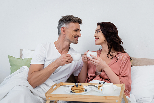 Smiling couple holding cups of coffee near blurred pancakes on tray in bedroom