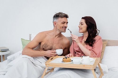 Cheerful woman in silk robe feeding husband with blueberry near breakfast on tray on bed