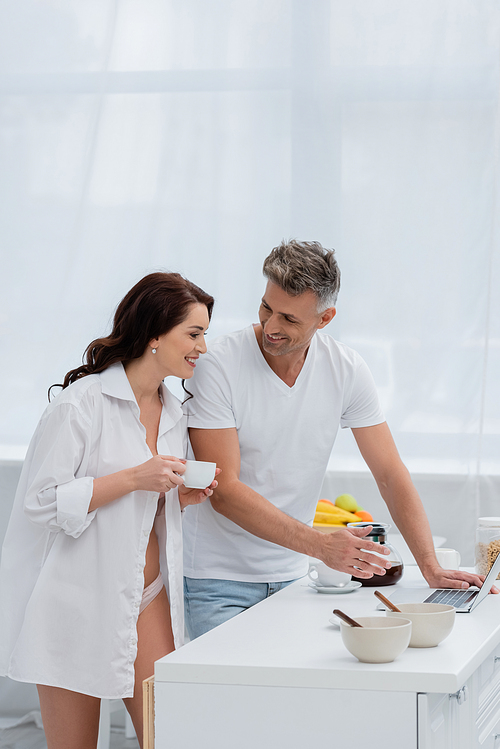 Smiling man pointing at laptop near sexy wife with coffee in kitchen