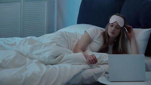 Woman in sleep mask looking at laptop on bedside table in bedroom