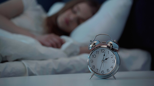 Alarm clock on bedside table near blurred woman sleeping on bed