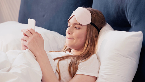 Young woman in sleep mask using smartphone on bed in morning