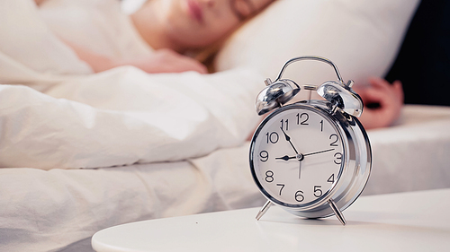 Cropped view of alarm clock on bedside table near blurred woman sleeping on bed