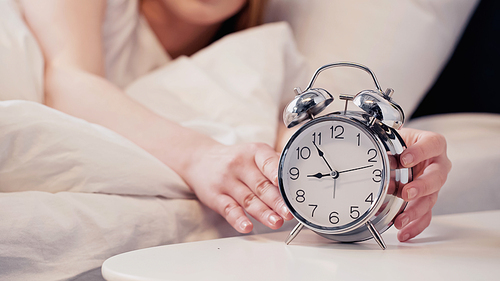 Cropped view of blurred woman touching alarm clock in bedroom in morning