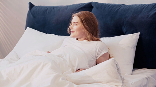 Positive young woman looking away while lying on bed in morning