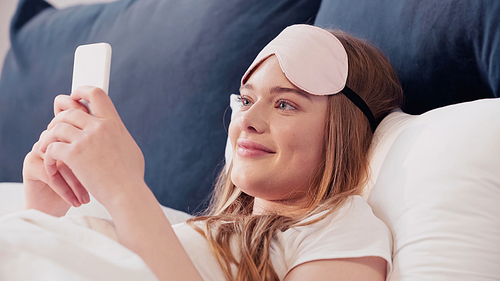 Cheerful woman in sleep mask using cellphone on bed at home