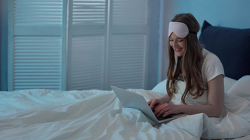 Cheerful woman in sleep mask using laptop on bed at night