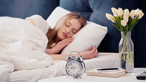 Alarm clock near smartphone and flowers on bedside table near blurred woman sleeping on bed