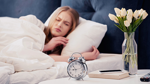 Displeased woman looking at alarm clock near smartphone in bedroom