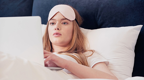 Freelancer in sleep mask using laptop on bed in morning