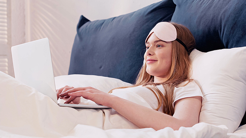 Positive freelancer in sleep mask using laptop while lying on bed in morning