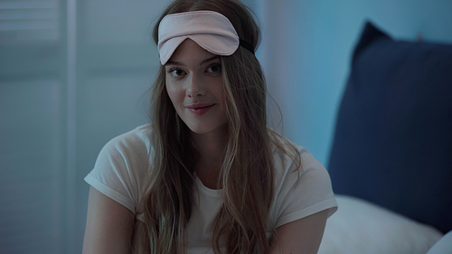 Smiling woman in sleep mask looking at camera in bedroom at night