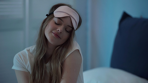 Young woman in sleep mask closing eyes while sitting on bed at night