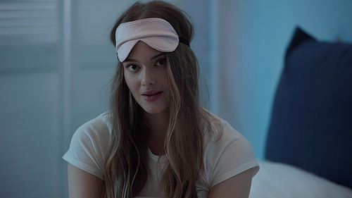 Blonde woman in sleep mask looking at camera in bedroom in evening