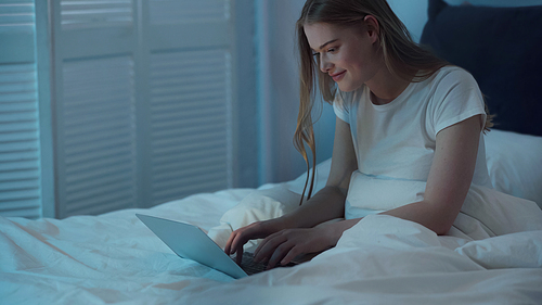 Cheerful woman using laptop while sitting on bed at night