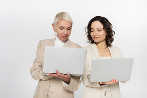 Senior businesswoman holding laptop near colleague isolated on grey