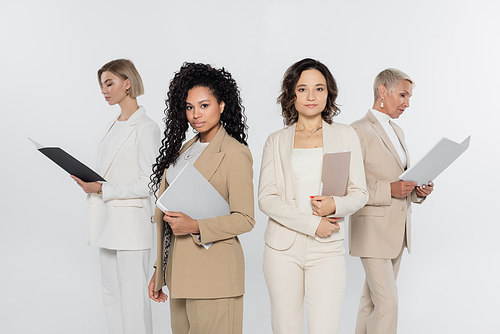 Interracial businesswomen with paper folders looking at camera near colleagues isolated on grey